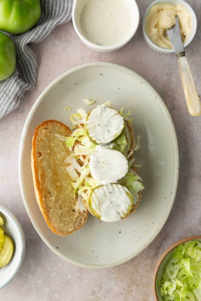 Open sandwich on an oval plate with fried green tomatoes, lettuce, pickles, and white sauce. Bread slice on the side. Surrounding items include green tomatoes, sauce, mayonnaise, and lettuce.