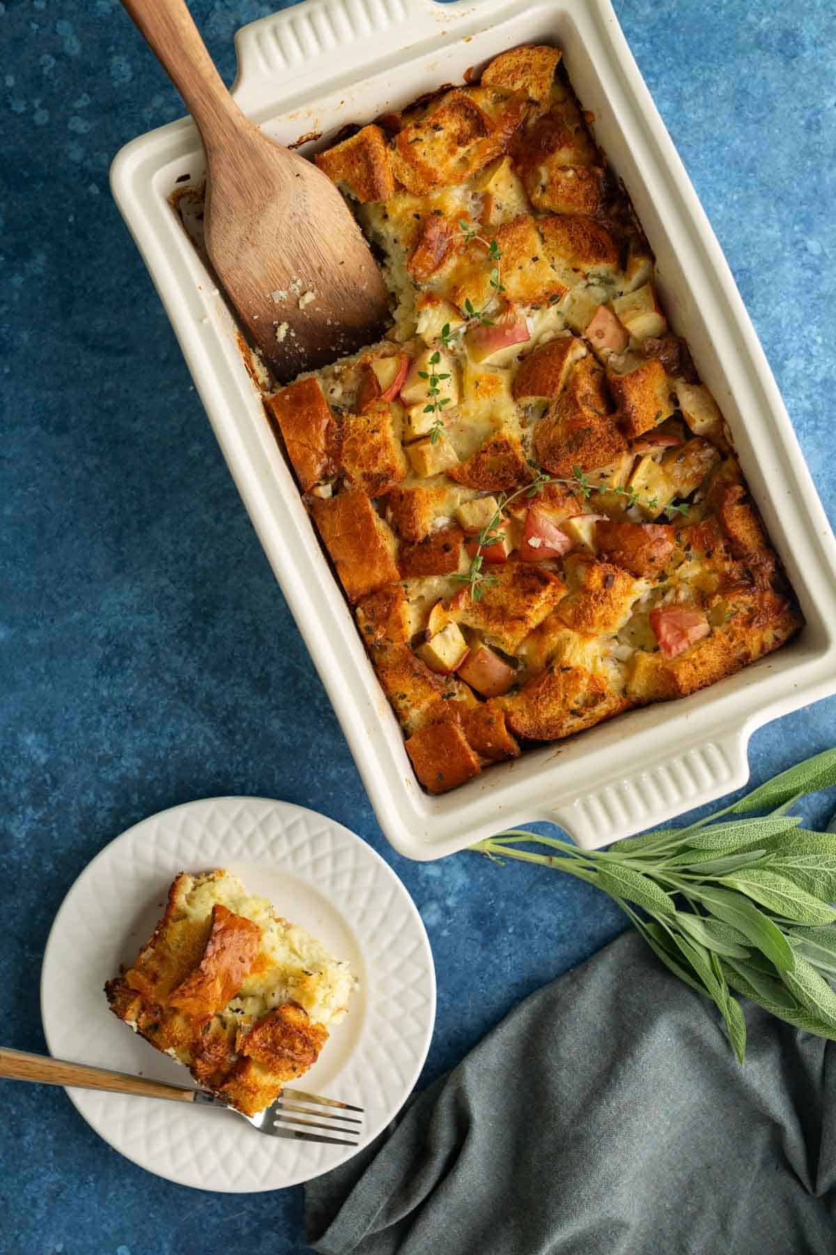A white baking dish with bread pudding topped with herbs, next to a plate holding a single serving, on a blue surface with sage leaves and a gray napkin.