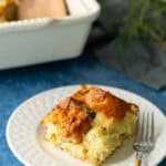 A square piece of breakfast casserole on a white plate with a fork, placed on a blue surface; casserole dish is in the background.
