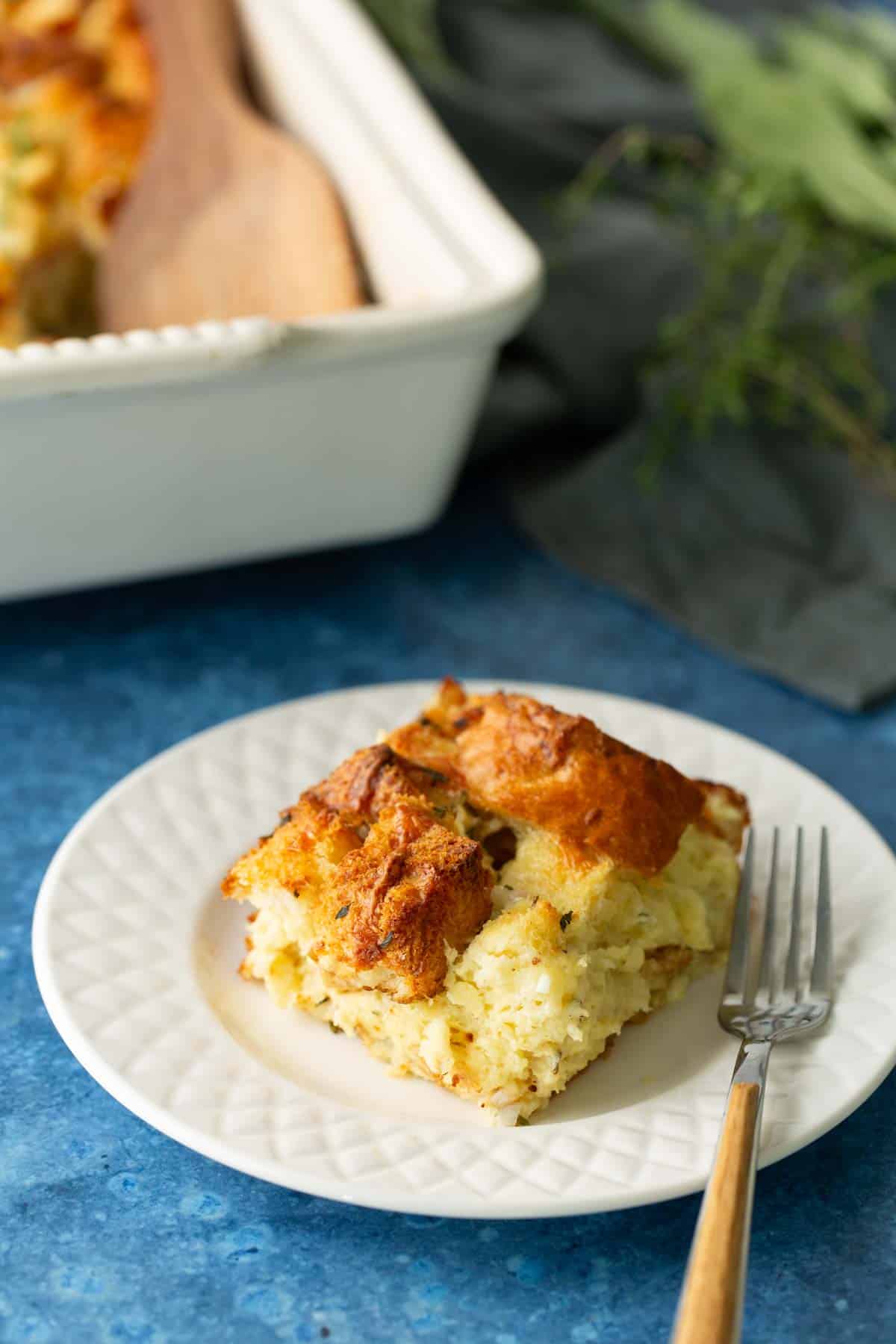 A square piece of breakfast casserole on a white plate with a fork, placed on a blue surface; casserole dish is in the background.