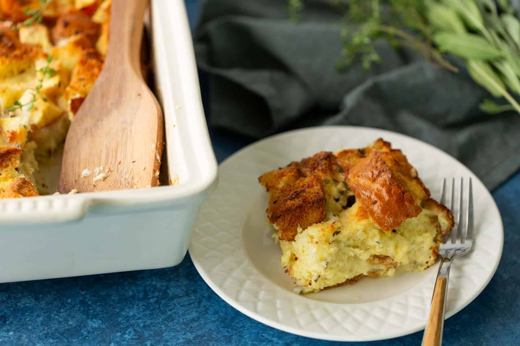 A slice of bread pudding is served on a white plate with a fork. The rest of the pudding is in a white baking dish with a wooden spatula. Herbs are in the background.