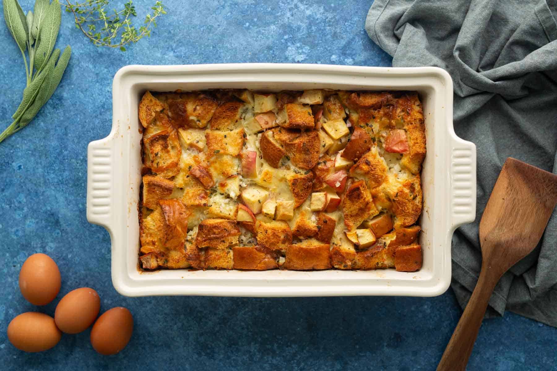 A baked casserole with cubed bread and apples in a white dish on a blue surface, surrounded by eggs, herbs, a wooden spatula, and a gray cloth.