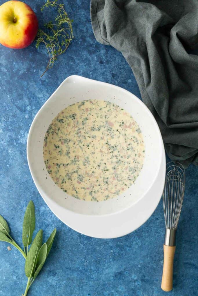 White mixing bowl with a creamy, speckled mixture on a blue surface. Nearby are a whisk, sage leaves, an apple, thyme, and a dark cloth.