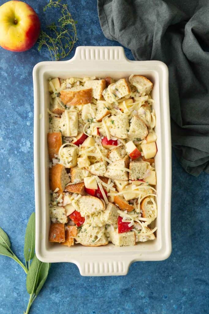 A rectangular baking dish filled with cubed bread, chopped apples, shredded cheese, and herbs sits on a blue surface with a green cloth and an apple nearby.