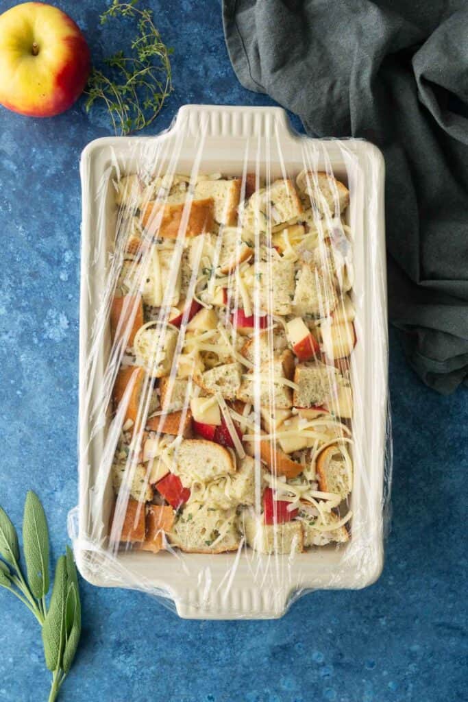 A casserole dish filled with bread cubes, apple pieces, and shredded cheese, covered with plastic wrap, sits on a blue surface with herbs and an apple nearby.