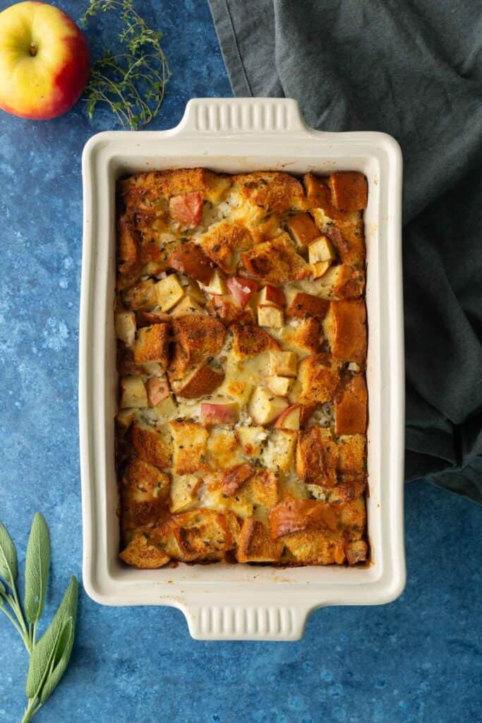 Rectangular white baking dish filled with cubed bread and apple stuffing, sitting on a blue surface with an apple, herbs, and dark cloth nearby.