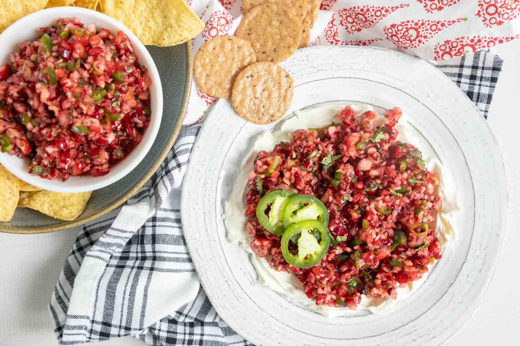Cranberry salsa served over cream cheese on a plate, with a bowl of salsa and tortilla chips nearby.