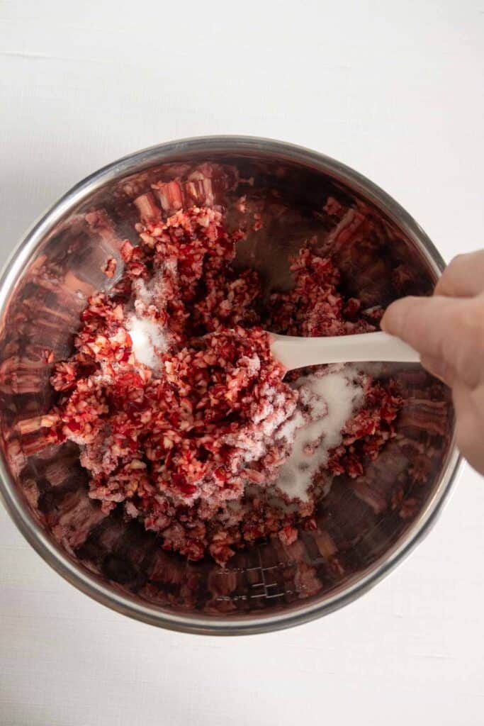A hand uses a white spatula to mix granulated sugar into a bowl of chopped cranberries.