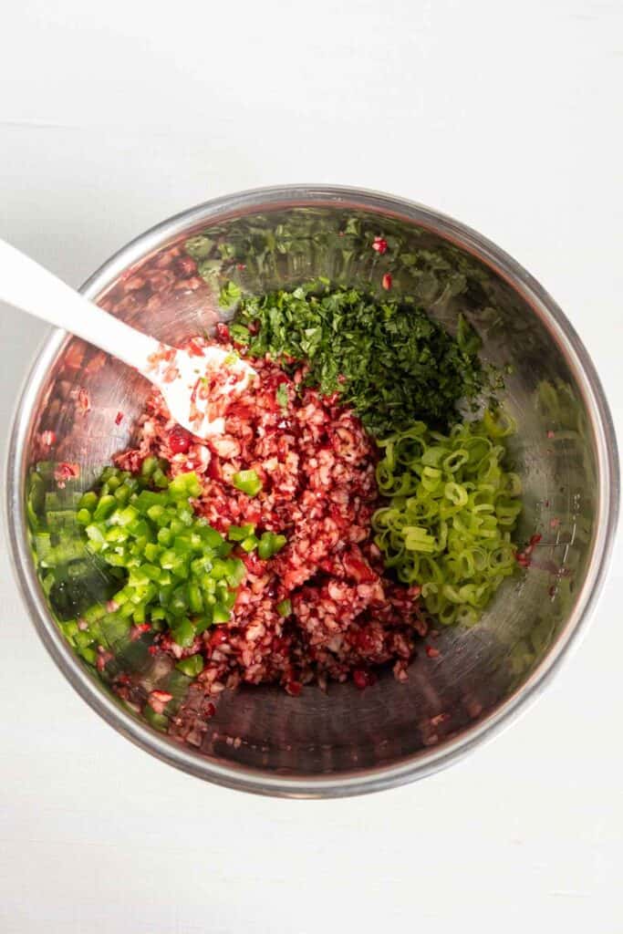 A metal mixing bowl containing chopped cranberries, jalapeños, cilantro, and green onions with a white silicone spatula.