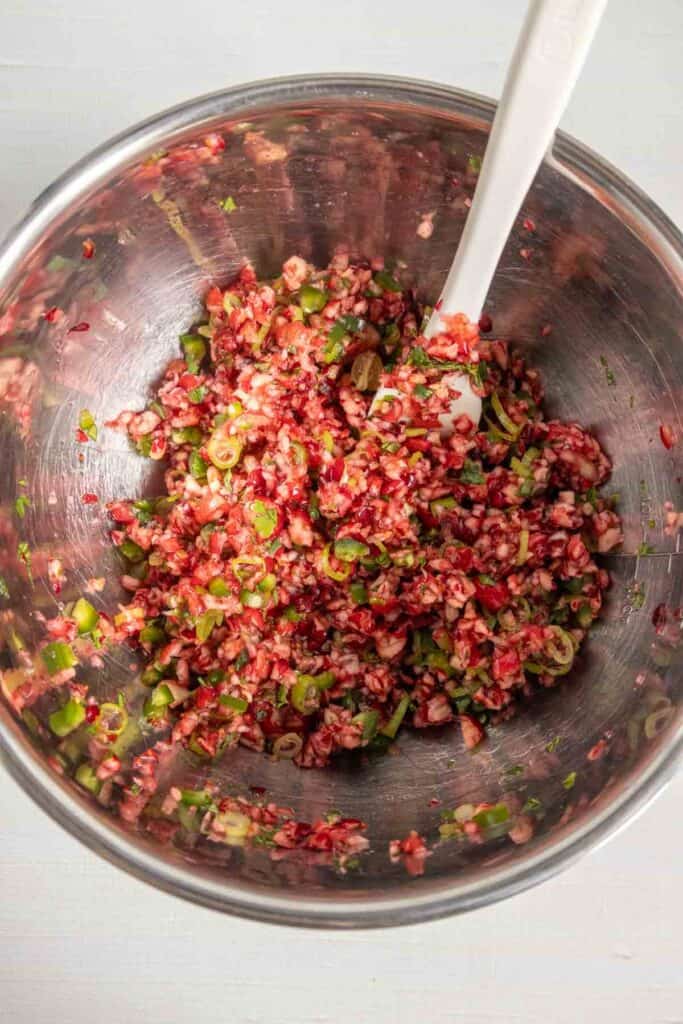 A metal mixing bowl containing a mixture of finely chopped cranberries, green peppers, and scallions with a white spatula.