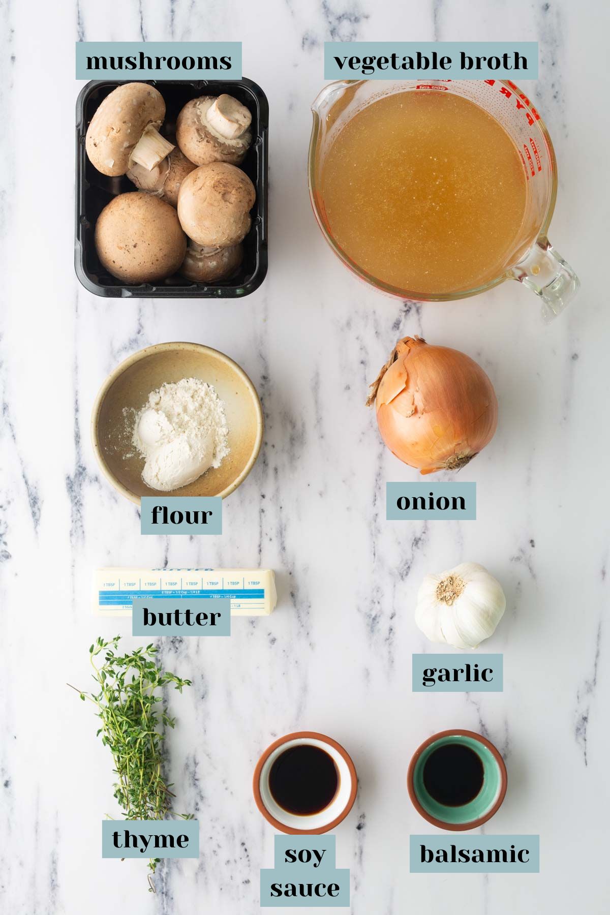 Ingredients for mushroom gravy arranged on a marble surface: mushrooms, vegetable broth, flour, an onion, butter, garlic, thyme, soy sauce, and balsamic vinegar.