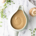 A ceramic gravy boat filled with mushroom sauce, surrounded by fresh mushrooms and sprigs of thyme on a marble surface.