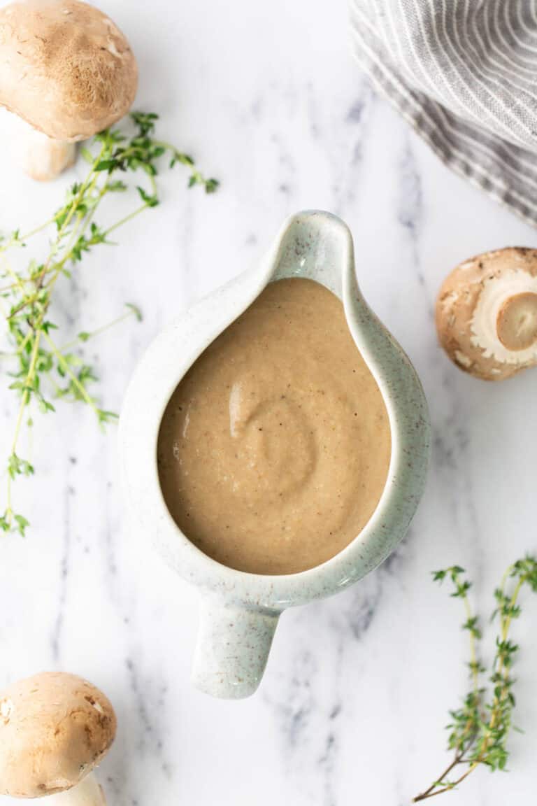 A ceramic gravy boat filled with mushroom sauce, surrounded by fresh mushrooms and sprigs of thyme on a marble surface.