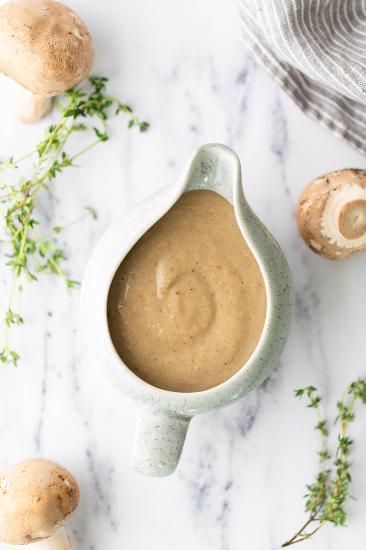 A ceramic gravy boat filled with mushroom sauce, surrounded by fresh mushrooms and sprigs of thyme on a marble surface.