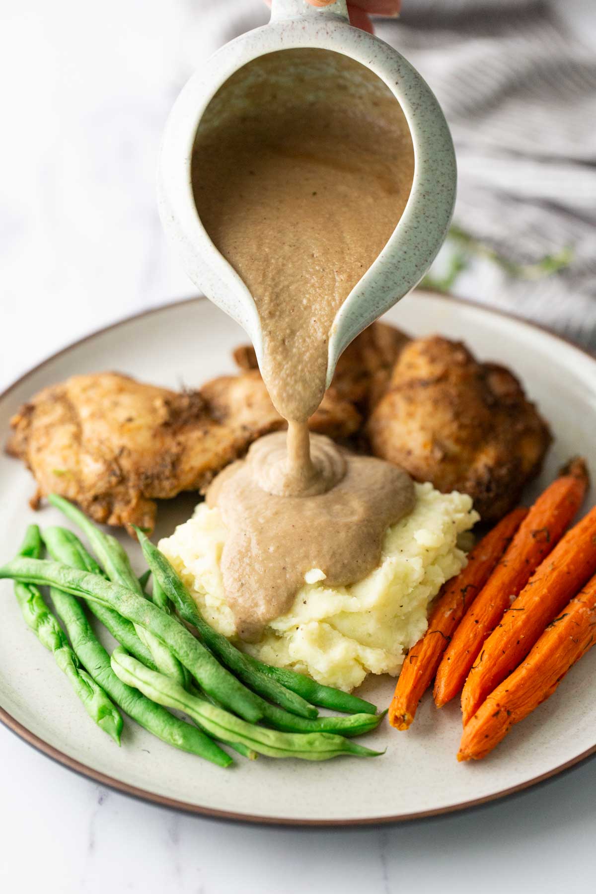 Brown gravy being poured from a pitcher onto a plate of mashed potatoes, roasted chicken, green beans, and carrots.