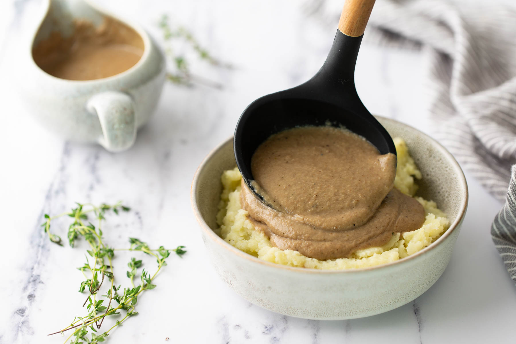 A black ladle pouring brown gravy over a bowl of mashed potatoes, with a small pitcher of gravy and fresh thyme sprigs nearby.
