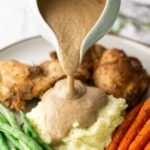 Creamy brown vegetarian gravy being poured from a ceramic pitcher onto mashed potatoes, served with roasted carrots and green beans.