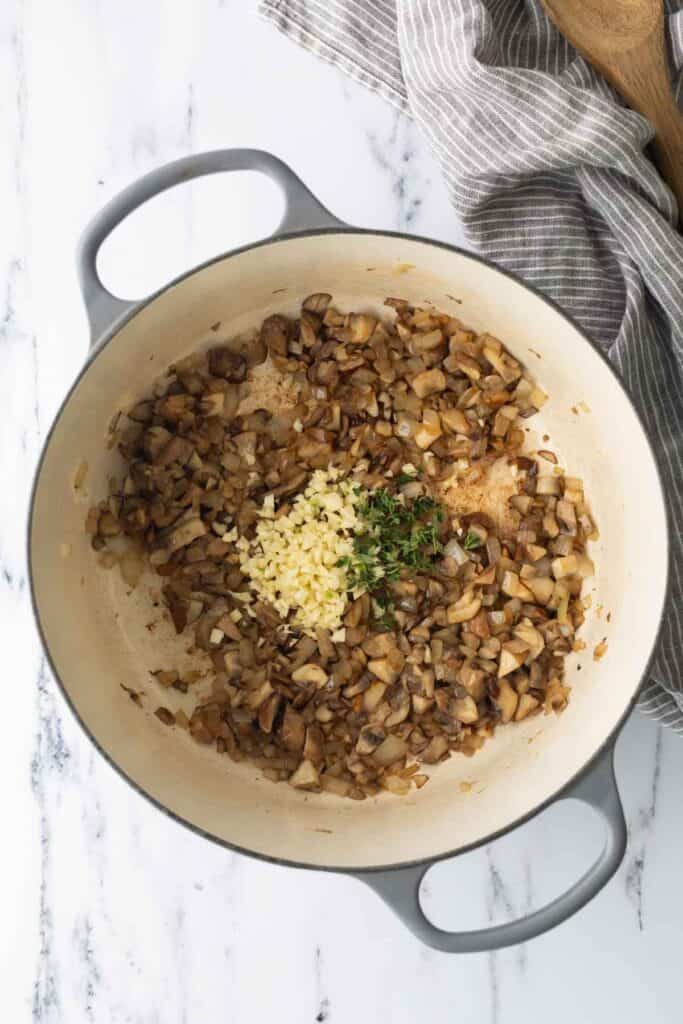 A Dutch oven containing cooked mushrooms and onions, topped with a mound of minced garlic and fresh thyme.
