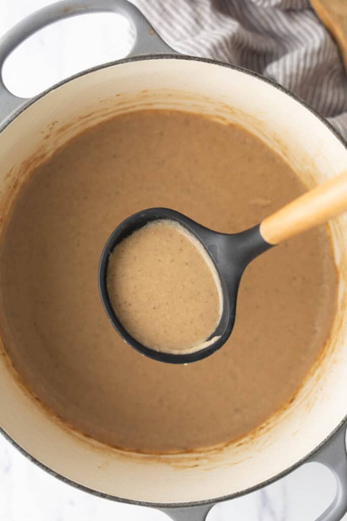 A ladle filled with creamy brown gravy held over a pot of the same sauce.