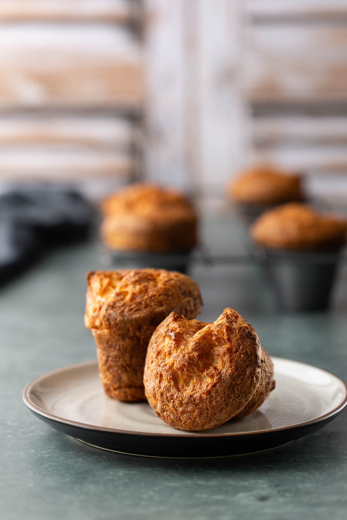 Two golden-brown popovers on a small plate, with more popovers in a muffin tin in the background.