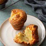 Two golden-brown popovers on a plate, one whole and one broken open to show the airy interior.