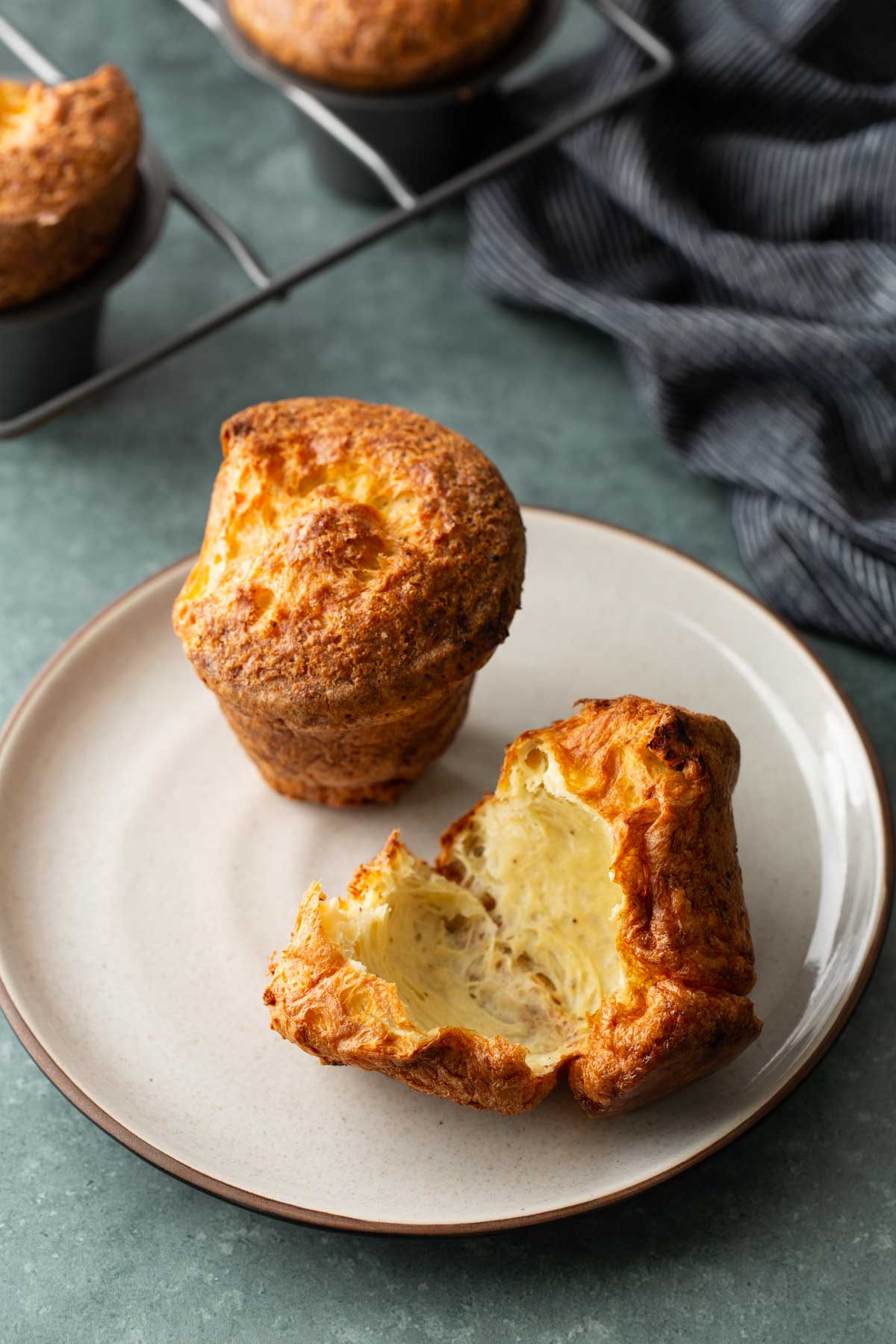Two golden-brown popovers on a plate, one whole and one broken open to show the airy interior.
