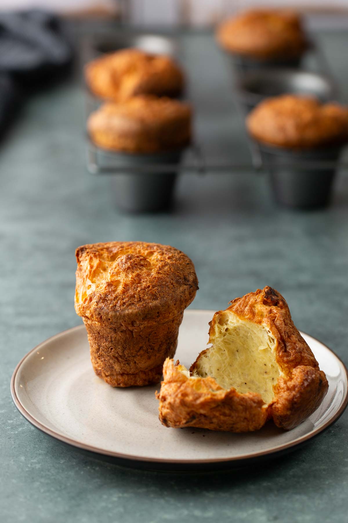 A whole popover and a halved popover on a plate, with additional popovers in a muffin tin in the background.