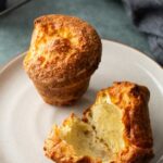 One whole golden-brown parmesan popover next to one that has been broken open on a plate.