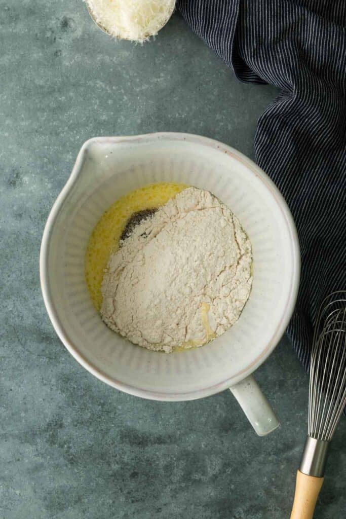 A ceramic mixing bowl containing flour, melted butter, and black pepper, with a whisk and a bowl of grated cheese nearby.