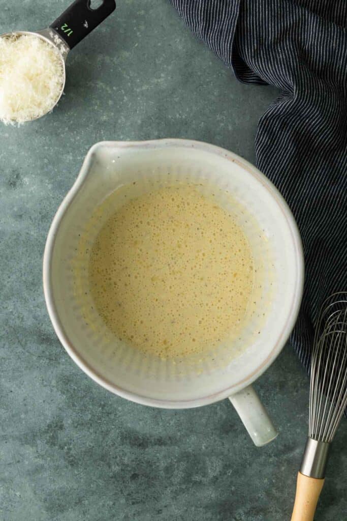A whisked egg and cheese mixture in a ceramic bowl, with a measuring cup of grated cheese and a wire whisk nearby.