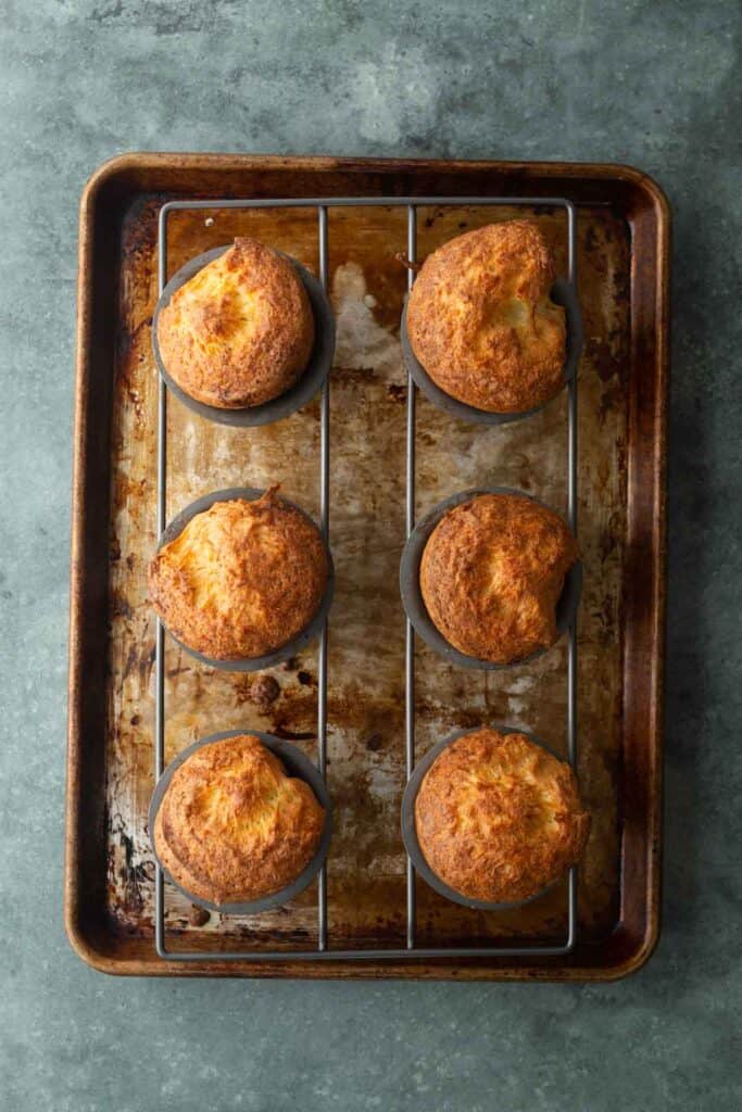 Six golden-brown popovers in a metal tin set on a baking sheet.