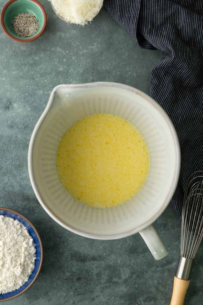 A ceramic mixing bowl containing whisked eggs, surrounded by bowls of flour, pepper, and cheese with a wire whisk nearby.