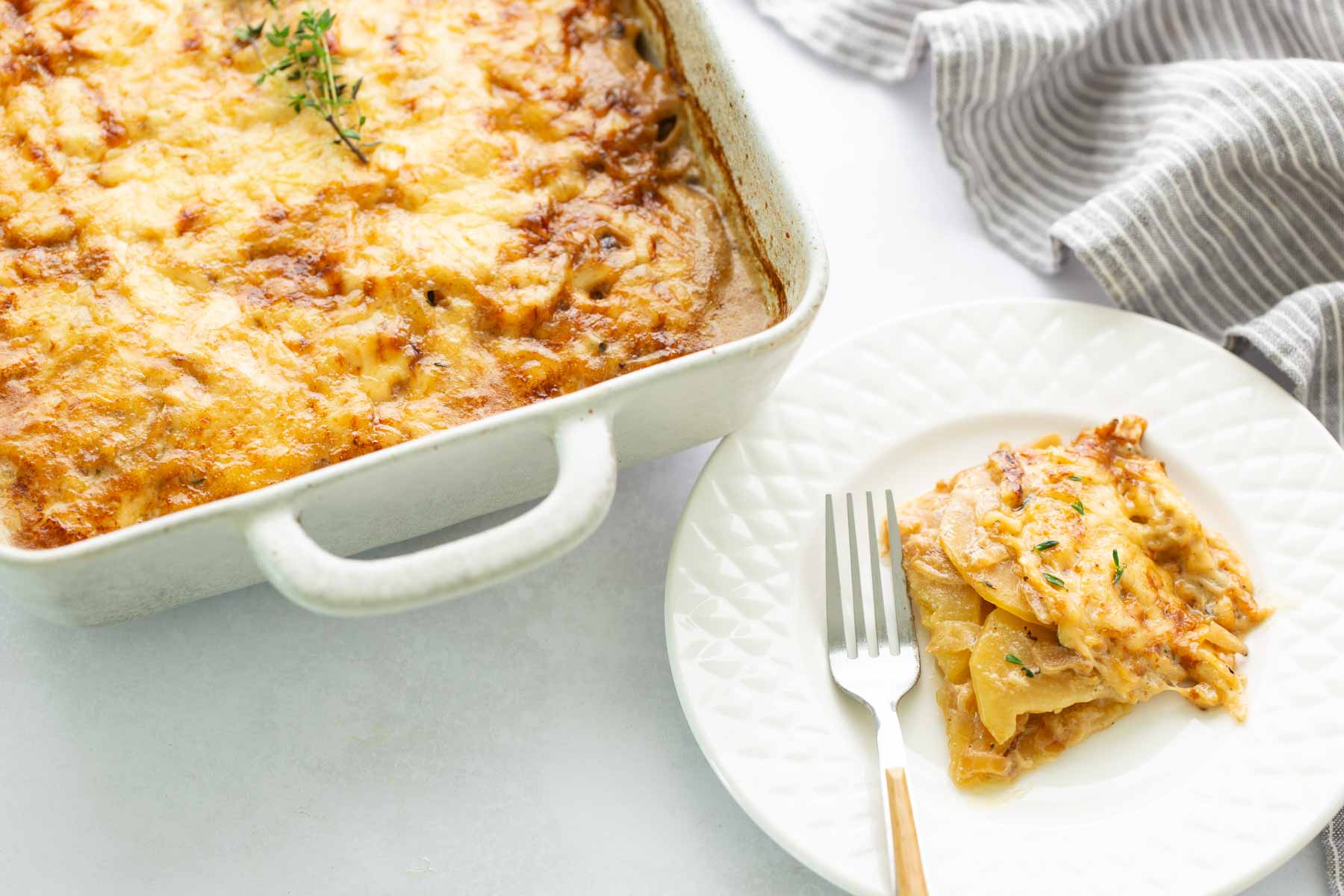 A baking dish of scalloped potatoes topped with melted cheese, with a single serving on a plate and a fork.