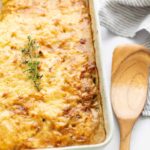A rectangular baking dish of cheesy scalloped potatoes topped with a sprig of fresh thyme, next to a wooden spatula.