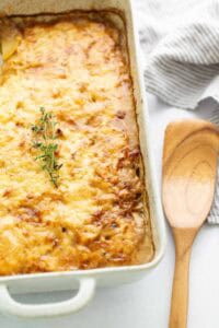 A rectangular baking dish of cheesy scalloped potatoes topped with a sprig of fresh thyme, next to a wooden spatula.