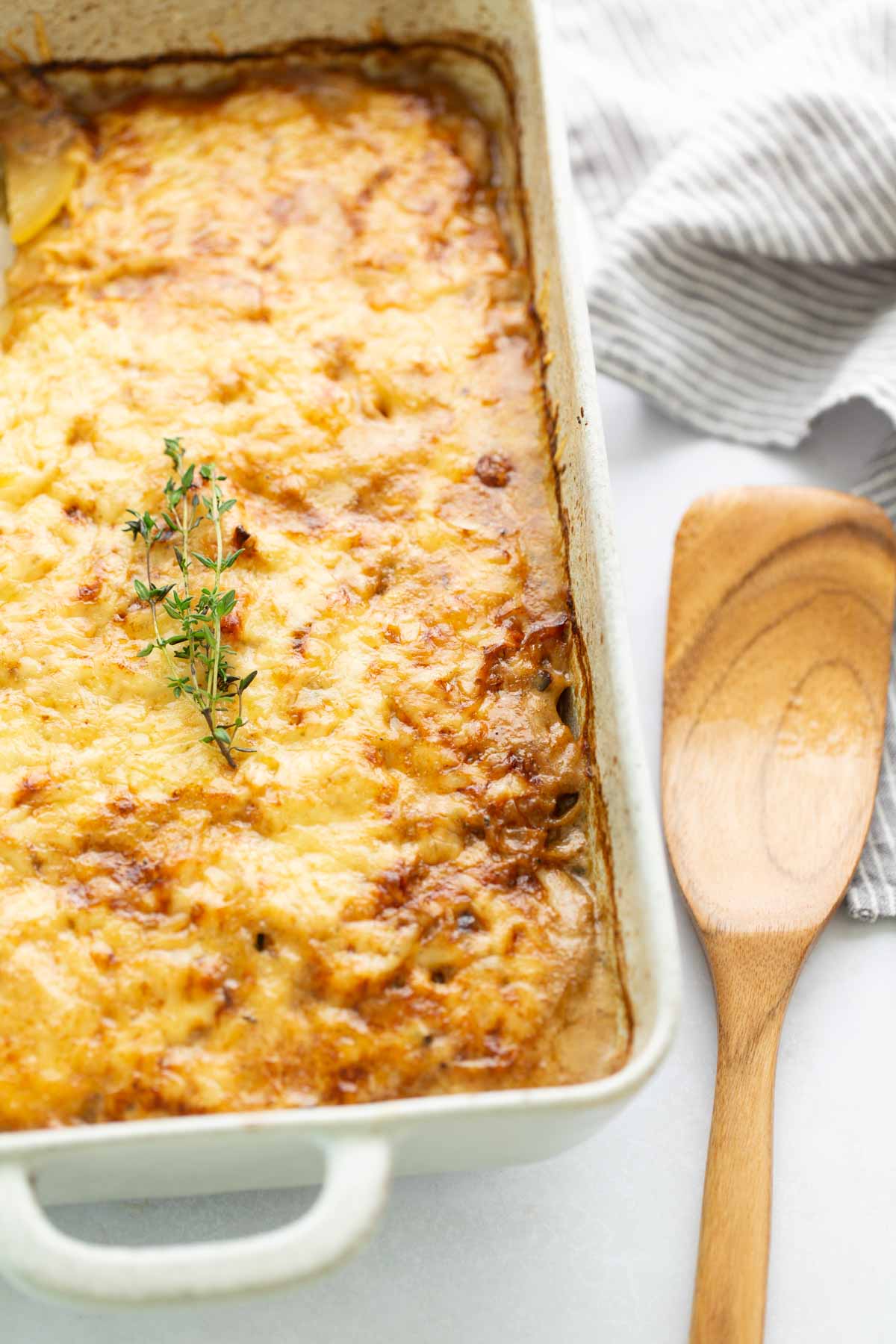 A rectangular baking dish of cheesy scalloped potatoes topped with a sprig of fresh thyme, next to a wooden spatula.