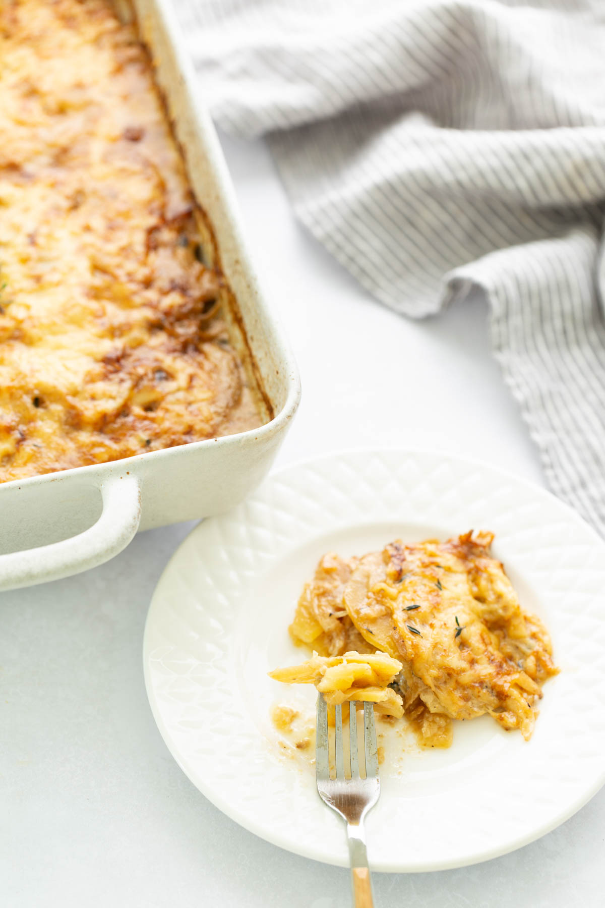 A serving of cheesy scalloped potatoes on a white plate with a fork, next to a baking dish of the same dish.