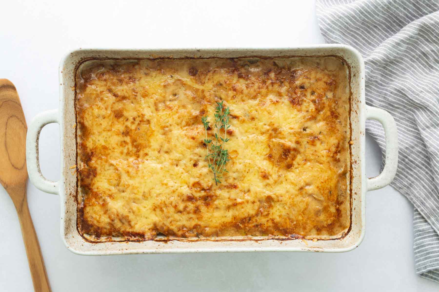 A rectangular baking dish containing a cheesy, browned casserole topped with a sprig of thyme.