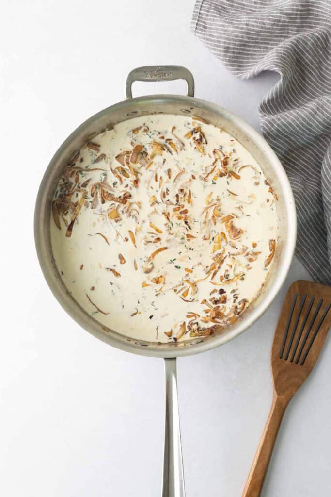 A stainless steel pan filled with a creamy sauce containing caramelized onions and herbs, with a wooden spatula nearby.
