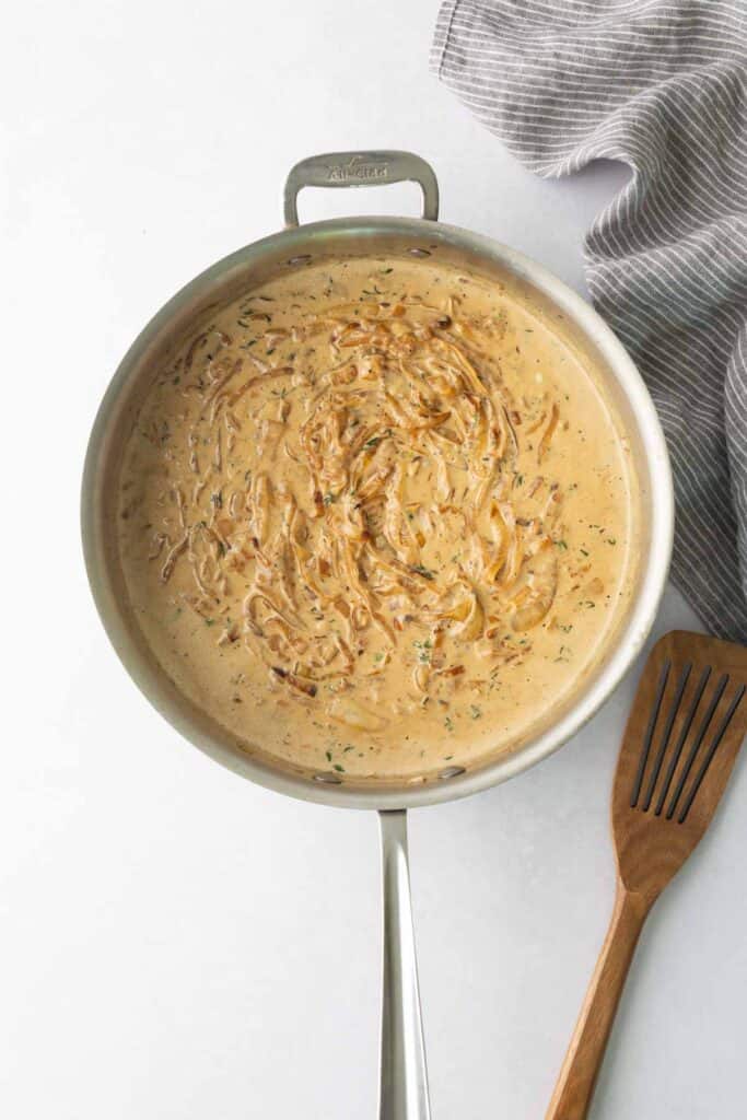 A creamy, onion-based sauce in a stainless steel skillet with a wooden spatula nearby.