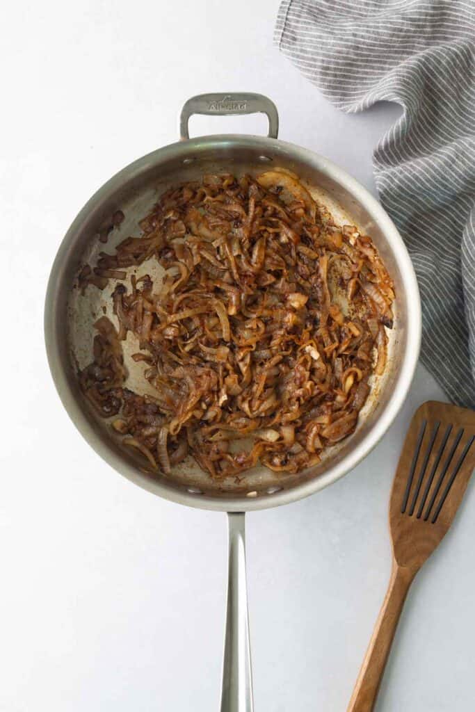 Caramelized onions in a stainless steel skillet with a wooden spatula and a grey striped cloth nearby.