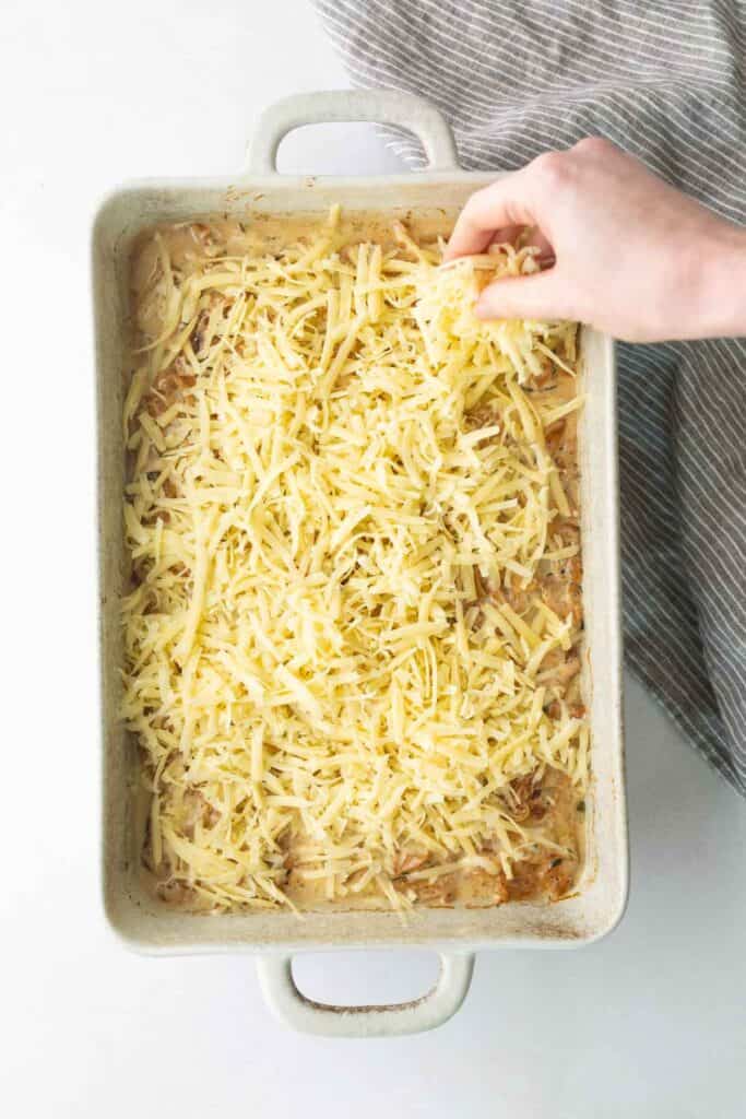 A hand sprinkles shredded cheese over a creamy casserole in a rectangular baking dish.